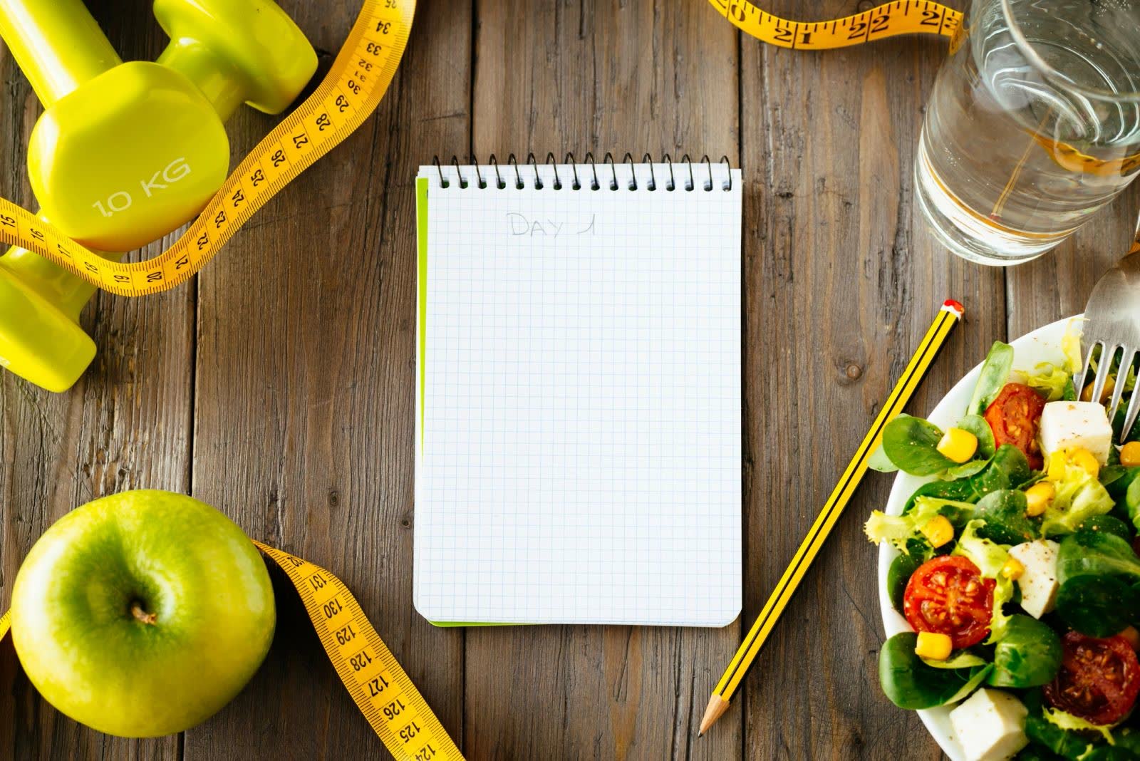 Dumbbells, an apple, a notepad, a tape measure and a salad on a wooden table