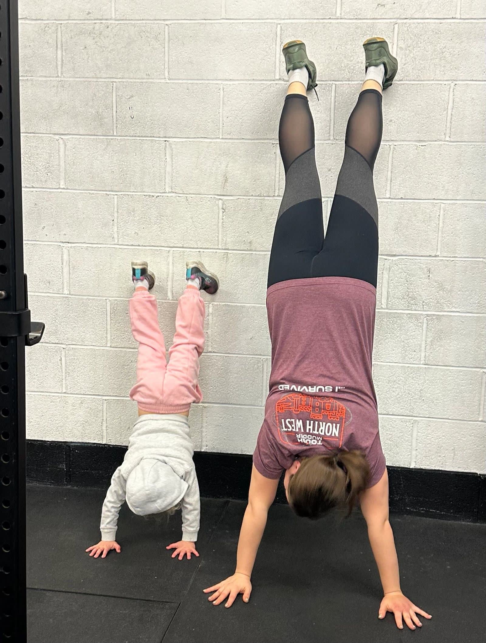 A mum and her young daughter doing handstands against the gym wall side by side