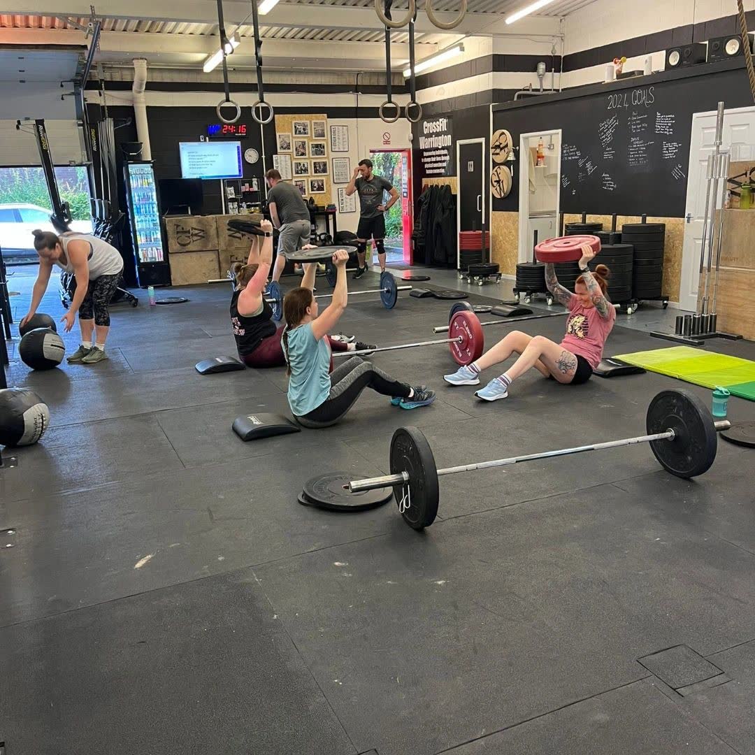 Members doing sit-ups on the gym floor, with loaded barbells laid out in front of them