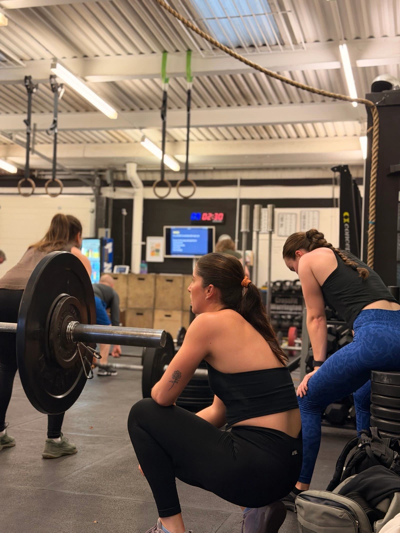 A member squatting beside a loaded barbell while others train around her