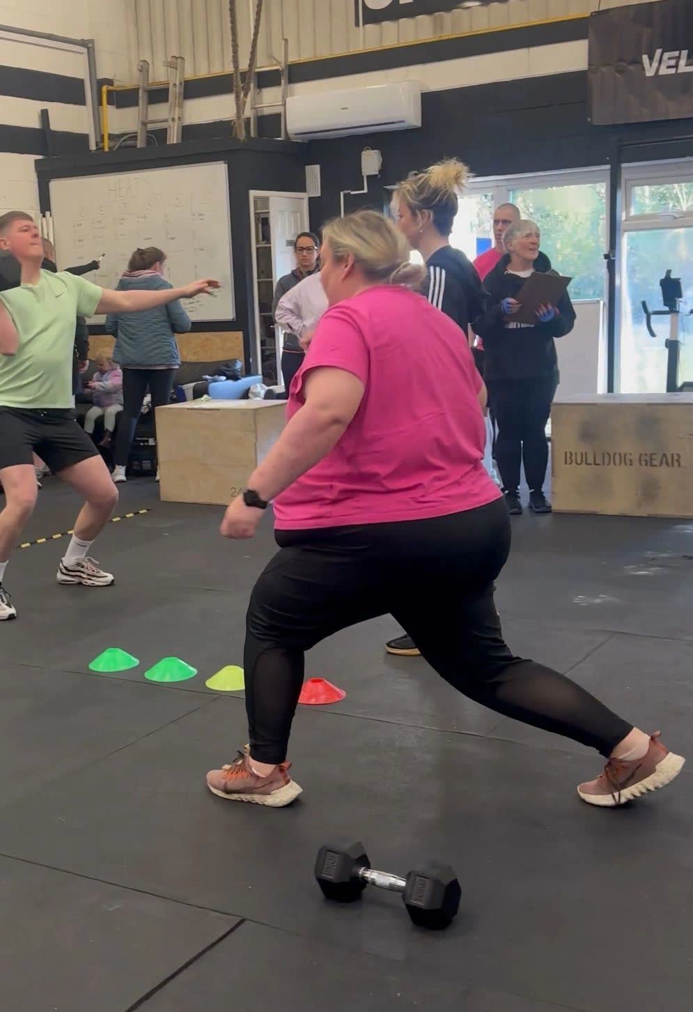 A member in a pink top lunging during a class, with cones and dumbbells on the floor