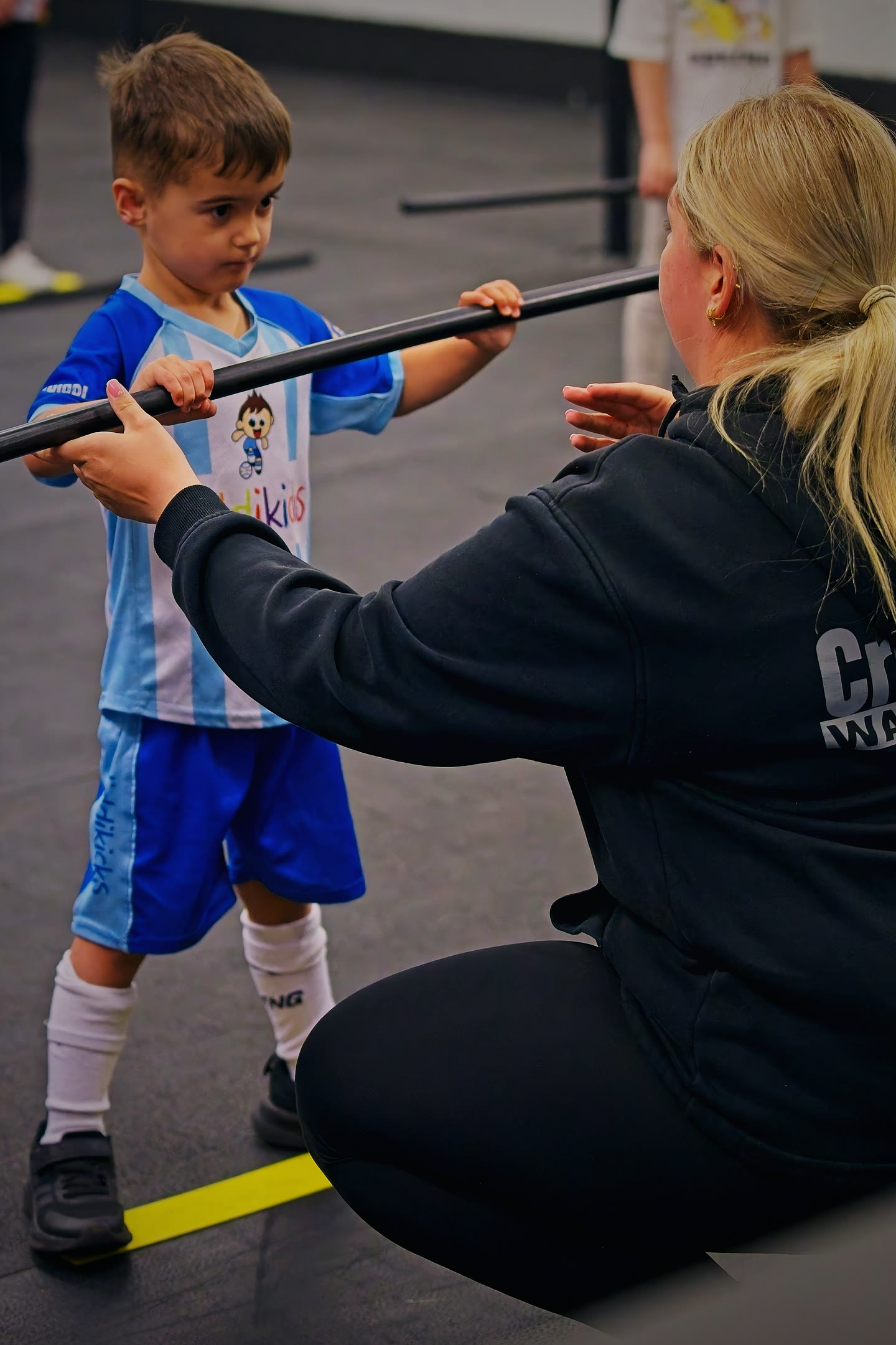 A coach helping a young boy hold a training bar at CrossFit Kids